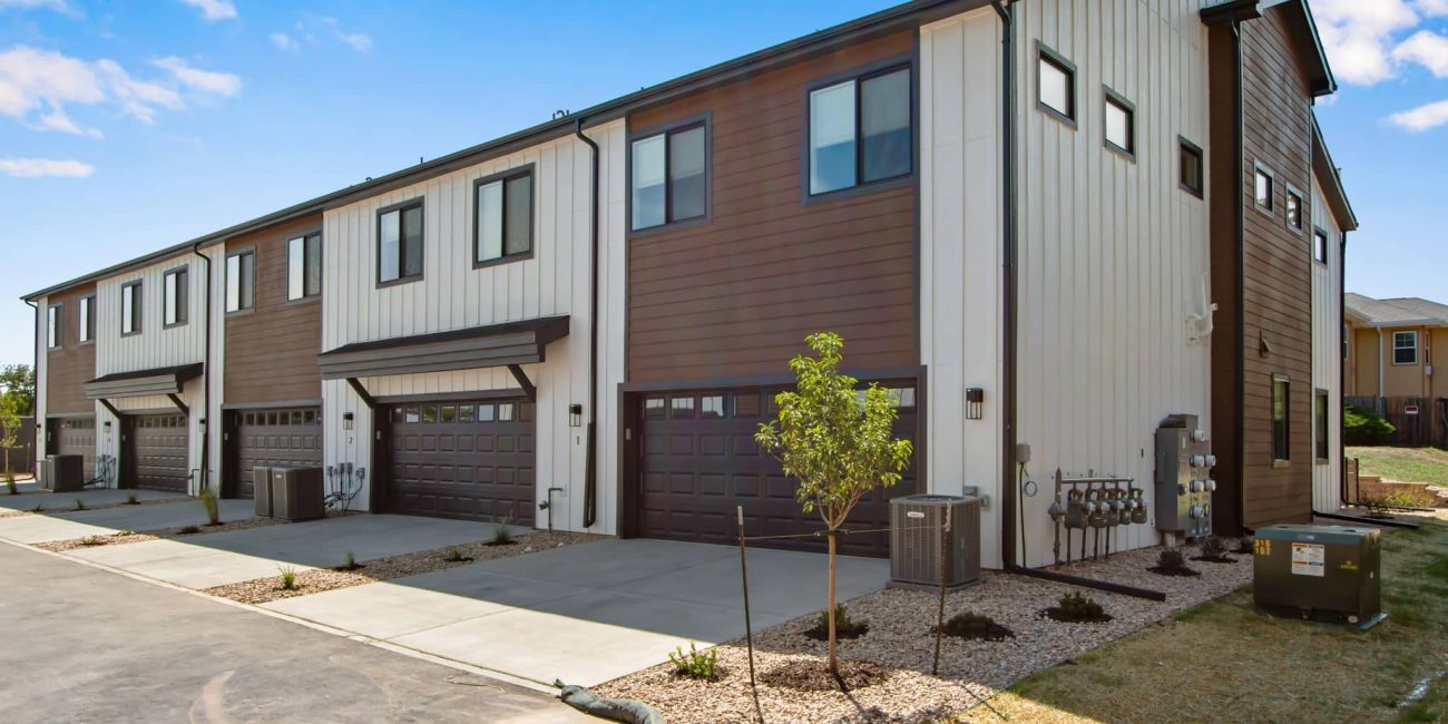 Rear view of Westlake Commons townhomes showcasing attached garages, clean lines, and a contemporary color palette of white and wood tones with paved driveways and minimal landscaping.